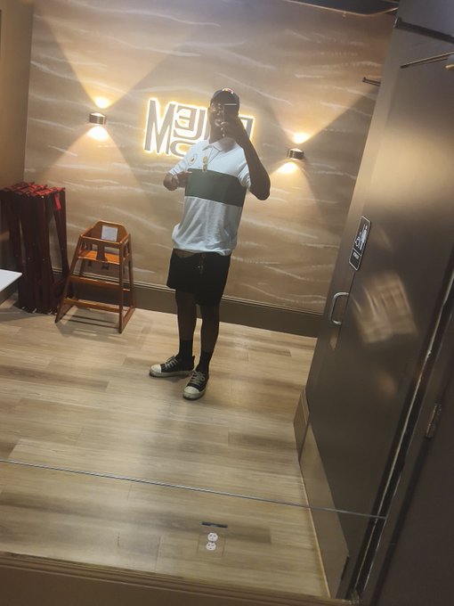 A mirror selfie of a man wearing a white and black striped shirt and black shorts, taken in a room with a wooden floor and a lit sign on the wall.
