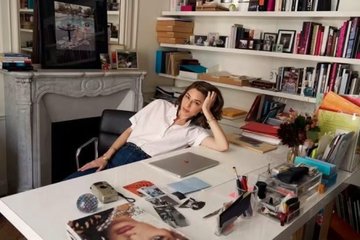 A woman sitting at a desk with a laptop, surrounded by books, photos, and art supplies in a cozy room with a fireplace, appearing relaxed or contemplative.