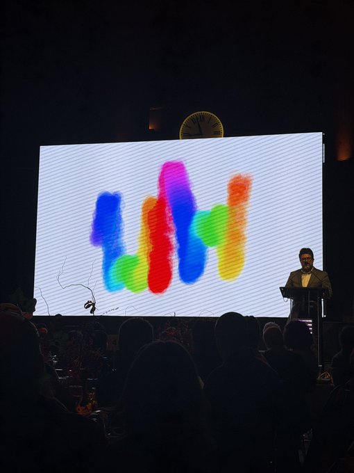 A person stands at a podium on a stage in front of a large screen displaying a colorful, hand-painted 'LYL' logo, with audience members visible in the foreground.