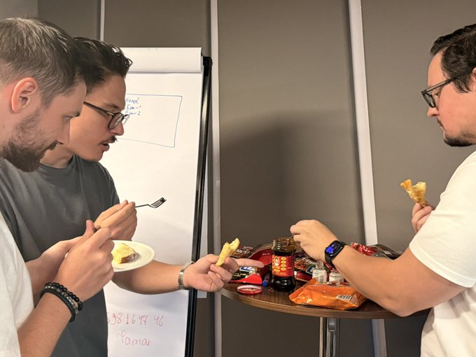 Three men at an indoor gathering sharing snacks from a small table, with one holding a fork and plate with cake, another eating a pastry, and a whiteboard with phone number '98 16 44 46' and 'fomar' visible in the background.