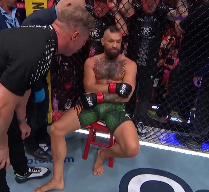 A UFC fighter with a beard and a large chest tattoo sits on a red stool inside the octagon, with his arms crossed and head down, while a referee or coach leans over him.