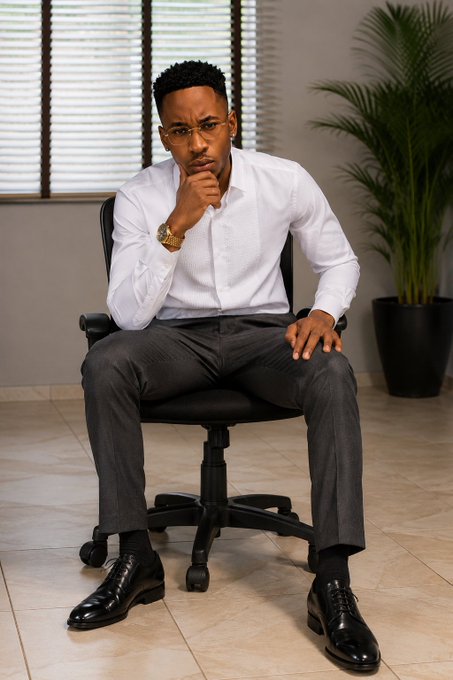 A well-dressed Black man with glasses sitting in a black office chair in a contemplative pose with his hand on his chin, wearing a white dress shirt, dark pinstriped trousers, black shoes, and a gold watch, with a potted palm plant in the background.