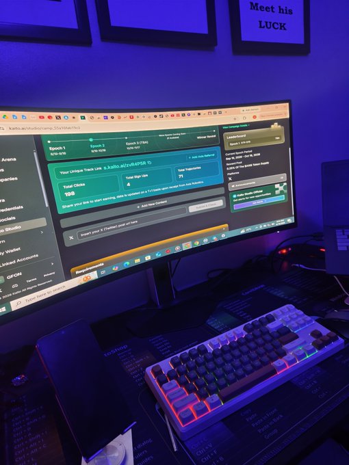 A dimly lit gaming setup with blue ambient lighting features a monitor displaying the website kaito.ai with tracking data, alongside a colorful RGB mechanical keyboard and mousepad with text like 'POSTING' and 'QUIET'.