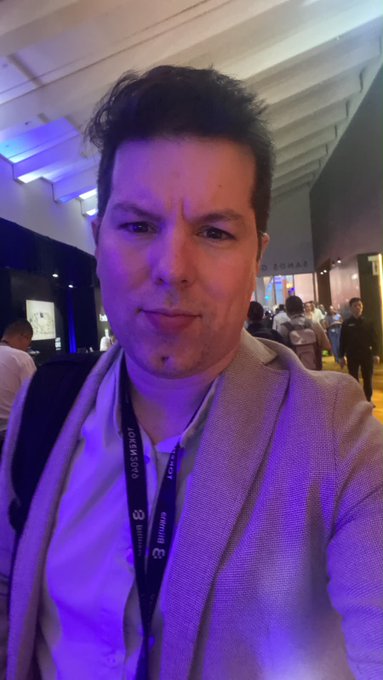 A selfie of a man wearing a light-colored blazer at what appears to be a conference, with a lanyard around his neck and blue lighting illuminating the background.