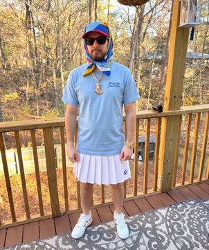 A man stands on a wooden deck wearing a light blue t-shirt with "Golfie" text, a white pleated skirt, a colorful cap, sunglasses, and a bandana around his neck, accessorized with a pendant necklace. The outfit humorously echoes the "Sorry for ruining NFTs" caption referencing Bored Ape Yacht Club, playfully blending casual golf attire with crypto culture parody.