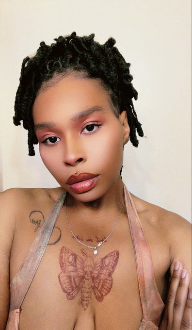 A woman with short dreadlocks and pink eyeshadow poses for the camera, featuring a large moth tattoo on her chest and a silver necklace.