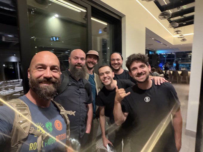 A group of six men pose together for a group selfie, smiling and flashing shaka hand signs in what appears to be a modern restaurant or lounge setting. One man's shirt reads "ROCK PAPER BITCOIN," tying into the post's theme of friends who share a passion for stacking Bitcoin (sats).