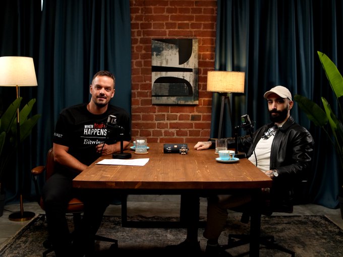 Two men sit at a wooden table in a podcast studio setting with a brick wall backdrop and dark blue curtains, recording an episode. The man on the left wears a black t-shirt reading "WHEN SHIT HAPPENS Bitwise," while the man on the right wears a beige cap and black jacket, suggesting this is the long-form conversation with Mert about the Zcash rumors mentioned in the post.