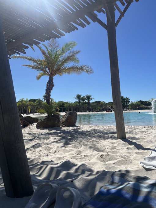 A sunny beach scene viewed from under a wooden pergola, showing white sand, a palm tree, a turquoise pool, and flip-flops on a towel in the foreground.
