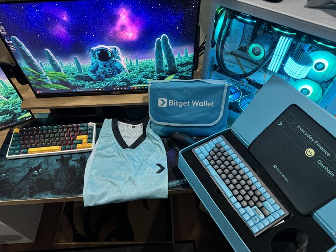 A Bitget Wallet branded keyboard rests on a desk alongside its matching cyan pouch and gift box, with a "Everyday Finance Onchain" insert, beneath a monitor displaying a cosmic astronaut wallpaper and a glowing custom PC. The setup showcases the exclusive keyboard promotional prize, tying into the post about testing the waves and offering the limited edition giveaway to followers.