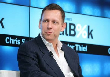 A man in a dark pinstripe suit and white shirt speaks at a 'Bloomberg' event, with text reading 'Chris S...' and '...er Thiel' visible on the screens behind him.