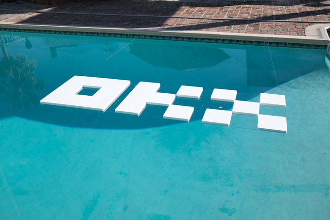 An outdoor swimming pool with the word 'OKX' spelled out in large white block letters floating on the water's surface.