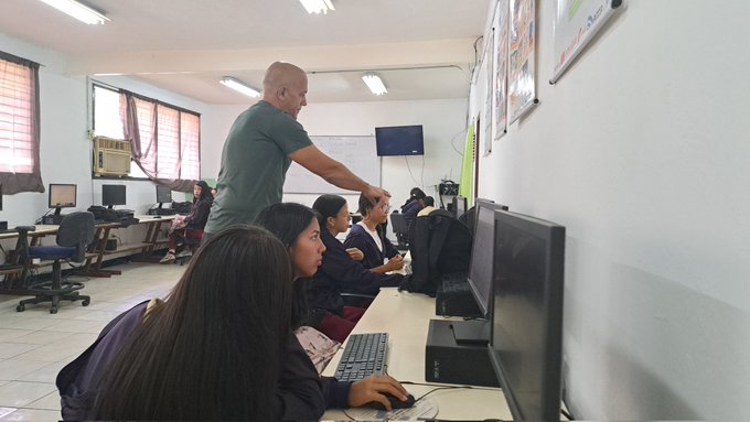 The image shows a group of people sitting at desks with computers, likely students or employees working together in an office setting. A man stands behind one of them, possibly giving instructions or guidance. The room has white walls and tiled floors, creating a professional atmosphere for learning or collaboration.