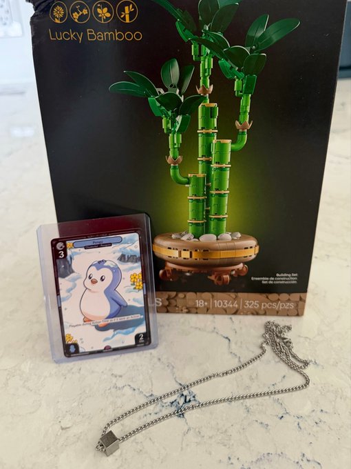 A LEGO "Lucky Bamboo" building set (10344, 325 pcs) sits on a marble surface alongside a Penguin TCG card and a silver chain necklace, tying into the "Pengu Promo" reveal with collectible and lifestyle flair.