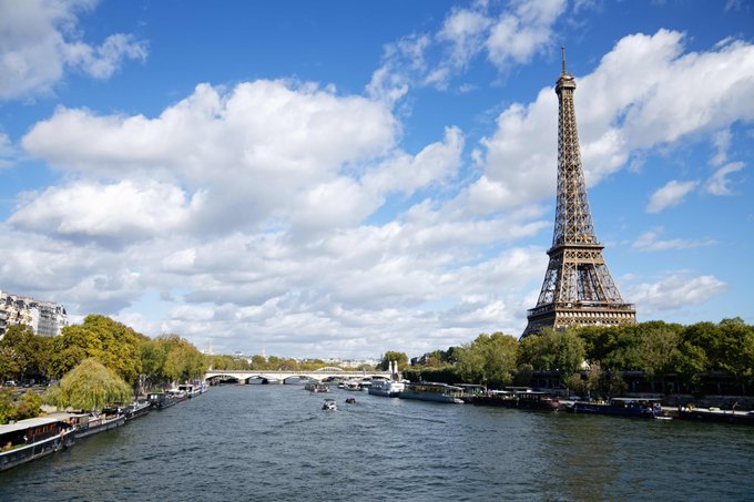 A daytime view of Paris shows the Eiffel Tower rising beside the Seine, with boats on the river, tree-lined embankments, bridges, and a blue sky filled with large clouds.