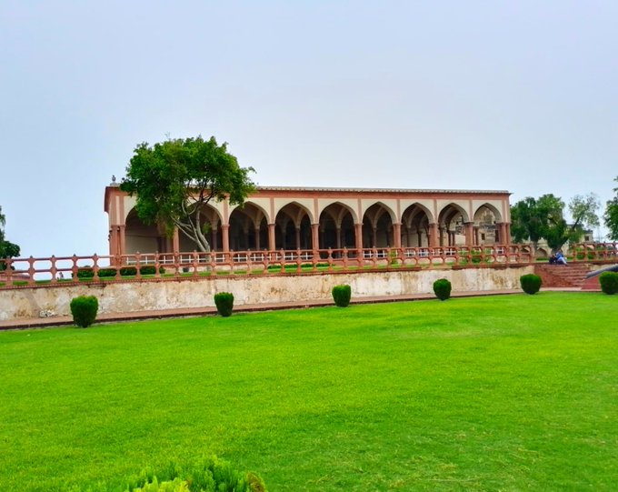 A historic pink-colored building with arched colonnades sits behind a manicured green lawn and stone balustrade, with a large tree to its left under an overcast sky.