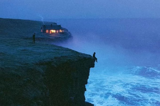 A small lit cottage sits atop a misty cliff while two solitary figures stand near the edge overlooking crashing ocean waves at dusk. The image evokes the quiet, contemplative mood of a fresh morning, perfectly complementing the serene "Good morning CT" greeting and the hopeful anticipation of a very good day ahead.