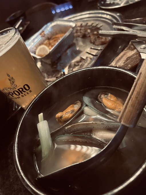 A close-up of a Korean BBQ-style meal featuring a Sapporo beer, grilled meat on a plate, and a steaming pot of mussel soup with chopsticks and green onions.