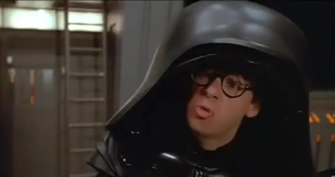 A man wearing round glasses and a black helmet and costume resembling Darth Vader from Star Wars speaks with a surprised expression.