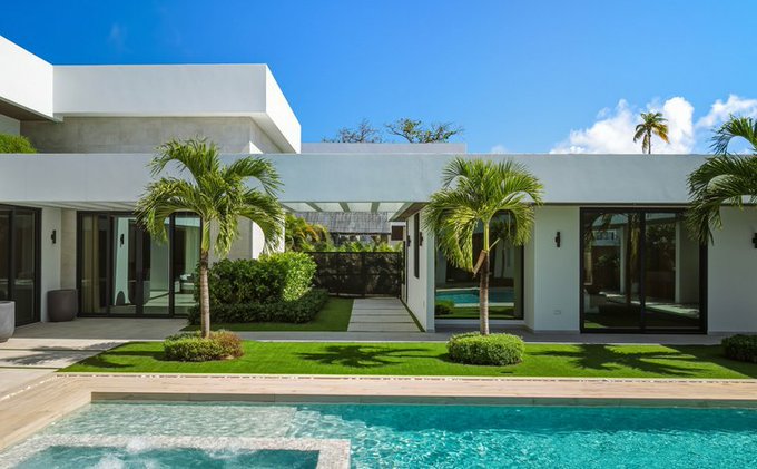 A modern white house with large glass windows, palm trees, a green lawn, and a swimming pool in the foreground under a blue sky.