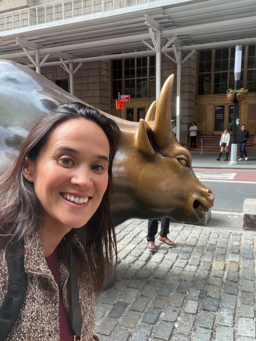A smiling woman posing for a selfie next to the Charging Bull statue on a cobblestone street, likely in New York's Financial District.