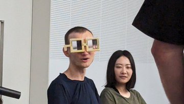 A photo of two people, with the person in the foreground wearing large pixel-style blocky glasses shaped like Minecraft tools, while a third person's arm is visible on the right.