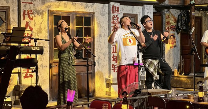 Three performers singing on stage with microphones, with Chinese characters displayed on the walls behind them.
