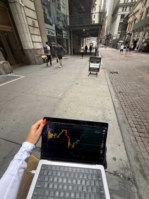 A first-person view of someone holding a laptop displaying a cryptocurrency trading chart while standing on a city street with pedestrians and buildings in the background.