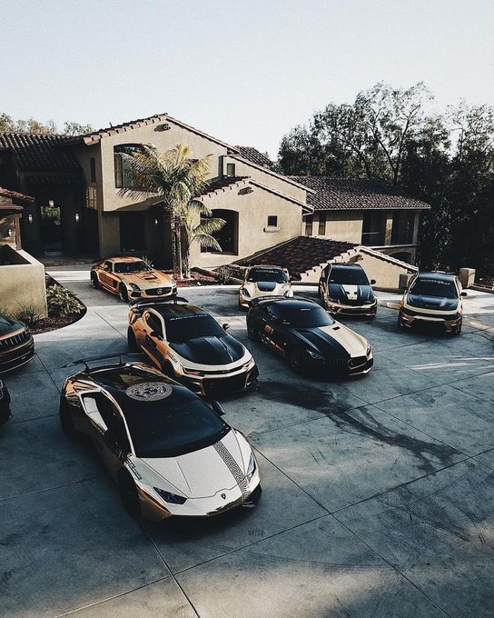 The image showcases a row of luxury sports cars parked next to each other, some in shades of black and gold. These high-end vehicles appear to be expensive and well-maintained, reflecting their owners' wealth and taste for fine automobiles. The cars are positioned on either side of the street in front of a large house or two-story home with a garage and patio. The image captures the attention of viewers as it appears that someone is taking a picture of these vehicles, adding an element of interest to the scene.