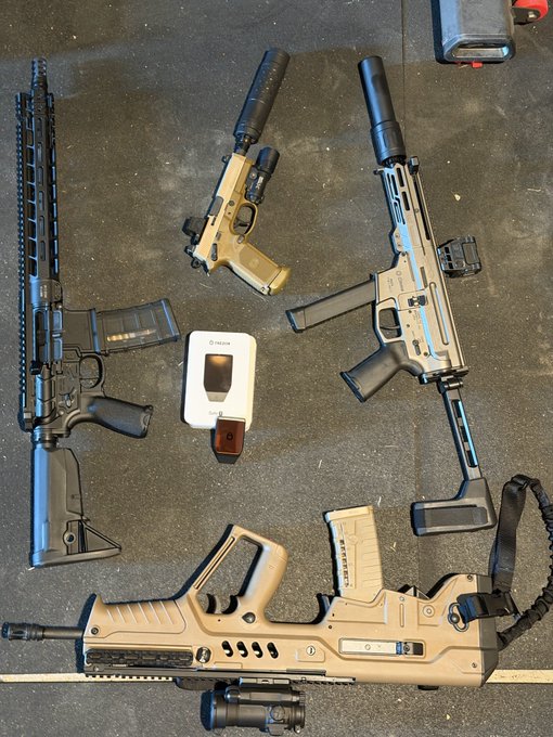 A collection of firearms—a tan Glock-style pistol, two rifles with suppressors and red dot sights, and a tactical shotgun—are laid out on a concrete floor alongside a Trezor hardware wallet, thanking the crypto brand for an excuse to visit the shooting range.