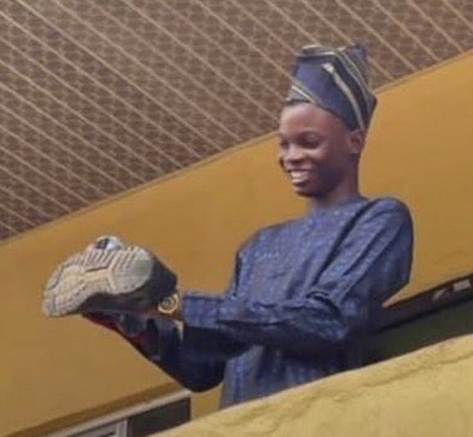 A young man wearing a dark blue traditional outfit and matching cap smiles while holding a packaged pair of sneakers.