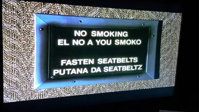 An illuminated sign with humorous 'Spanglish' text reads: 'NO SMOKING EL NO A YOU SMOKO' and 'FASTEN SEATBELTS PUTANA DA SEATBELTZ'.