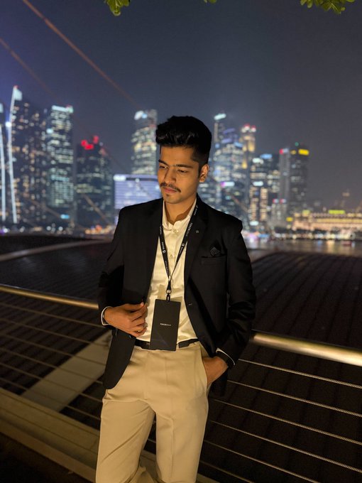 Portrait of a young man in a black blazer and beige pants with a lanyard, posing at night with a city skyline of illuminated skyscrapers in the background.