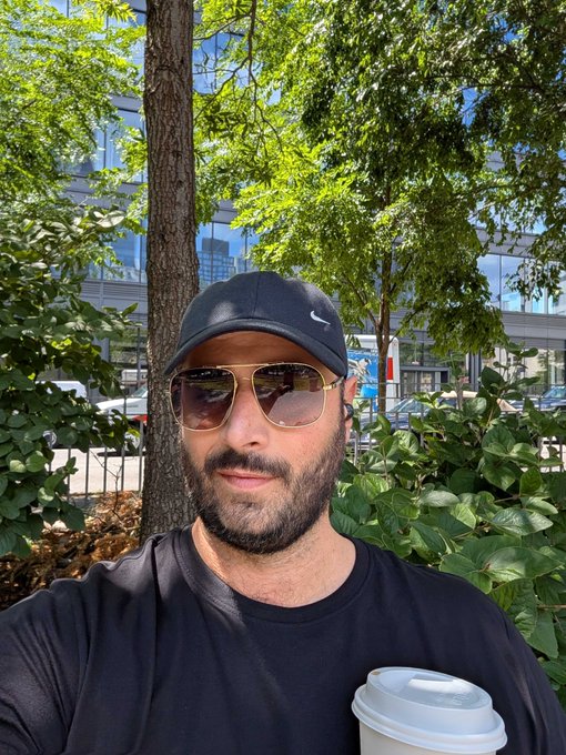 The image features a man wearing sunglasses and a black t-shirt holding a coffee cup while standing outside, possibly in New York City. The photo appears to be taken by someone who wants to share their experience with friends or followers on social media platforms.