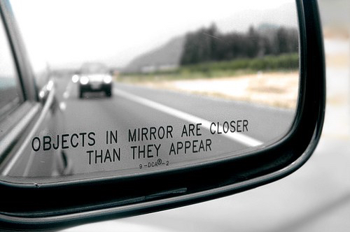 A view through a car's side mirror showing a road and another vehicle, displaying the standard warning text "OBJECTS IN MIRROR ARE CLOSER THAN THEY APPEAR."