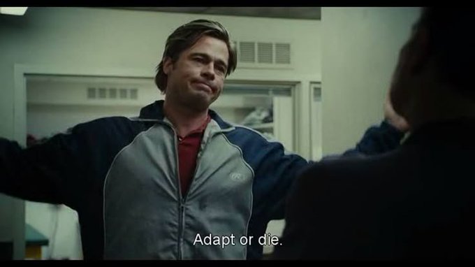 A still from a film showing actor Brad Pitt in a gray and blue track jacket, with the subtitle "Adapt or die." at the bottom.
