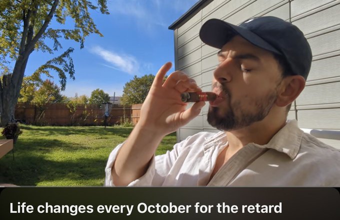 A man wearing a black cap takes a hit from a cannabis vape pen in a sunny backyard, with the caption "Life changes every October for the retard" displayed below, humorously teasing the post's reference to the changing seasons by suggesting October brings personal transformation.