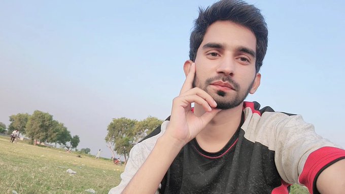 A young man takes a selfie outdoors in a grassy park, resting his hand on his face with a thoughtful expression under a clear sky.