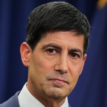 A close-up portrait of a middle-aged man with dark hair wearing a suit and tie, with a dark blue background, appearing to be in a formal setting.