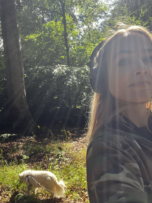 A selfie of a woman in a camo hoodie with a white dog on a leash in a sunlit wooded area.