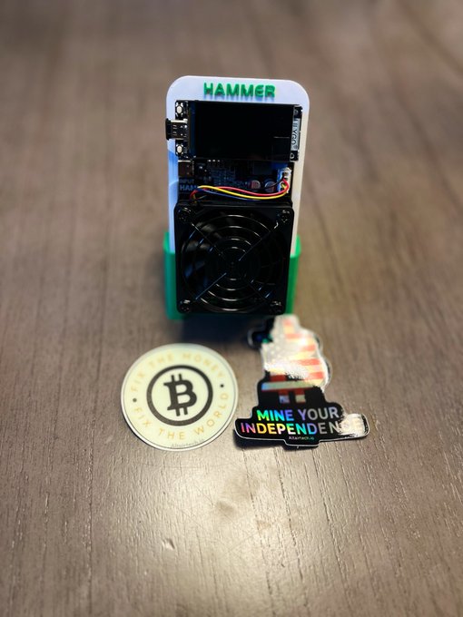 A Hammerminer BC01 Bitcoin mining device labeled "HAMMER" sits on a wooden surface alongside a "Digital Money for the World" Bitcoin coin and a colorful sticker reading "MINE YOUR INDEPENDENCE," showcasing the prize awarded to an Instagram giveaway winner.
