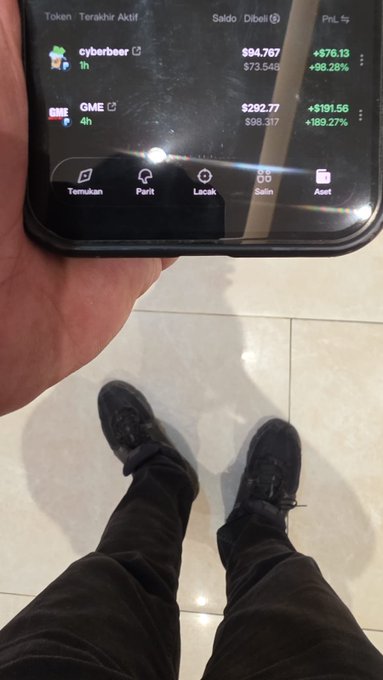 A person holds a phone displaying a crypto/stock app showing 'cyberbeer' at +98.28% and 'GME' at +189.27%, with their feet visible on a tiled floor.