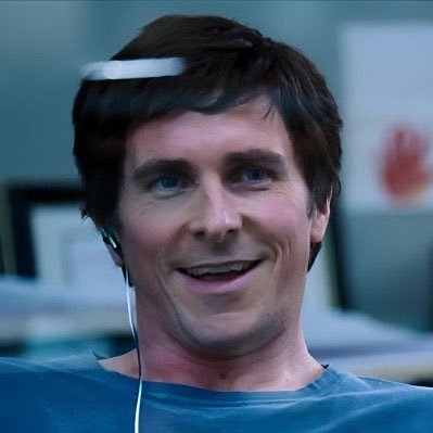 A smiling man wearing a futuristic thin white headband device across his forehead and white earbuds looks off-camera in what appears to be an office setting.