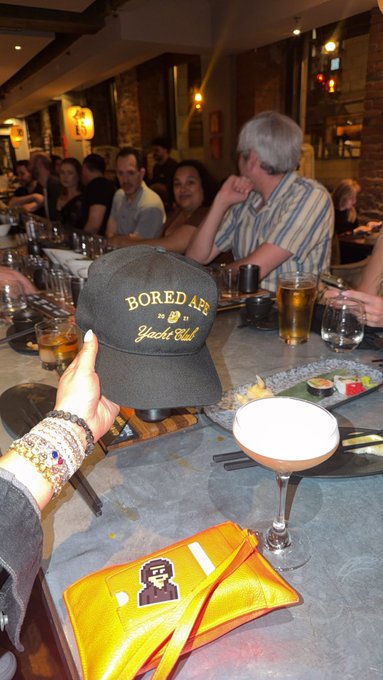 The image shows a group of people sitting around a table at a restaurant, with some of them wearing hats that say "bored ape yacht club". The scene appears to be set for a casual gathering or party, as there are various drinks on the table, including wine glasses and cups filled with beer. A person is holding up an orange wallet in front of the group, possibly showing off their new purchase or sharing some interesting information about it.