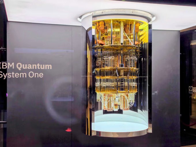 A glowing cylindrical IBM Quantum System One is displayed behind glass with the "IBM Quantum System One" label visible on the adjacent wall. The image of advanced quantum computing hardware visually alludes to the cryptographic security concerns central to the Bitcoin Security Consortium's $15 million pledge by BlackRock, Coinbase, Strategy, and Anchorage Digital, as quantum computing poses potential future threats to Bitcoin's cryptographic foundations.