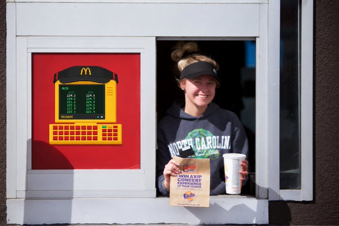 A smiling worker at a McDonald's drive-thru window hands out a drink and a paper bag promoting a contest, with a digital menu board visible on the left.
