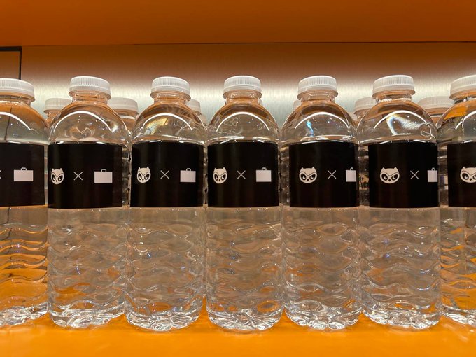 A row of clear water bottles with black labels featuring a small owl icon, an "x," and a briefcase graphic are neatly arranged on an orange shelf, tying into the "Moonbirds x StonkBrokers" collaboration by blending the Moonbirds owl mascot with StonkBrokers' signature briefcase branding.