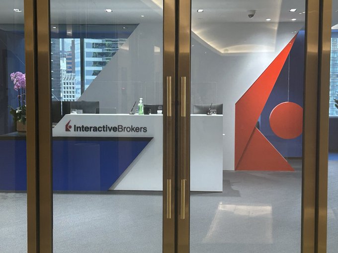 A view into the Interactive Brokers office lobby, featuring a reception desk with the "Interactive Brokers" logo and a large red and blue triangular wall design. The image relates to the post by illustrating the physical Interactive Brokers office, likely referencing the "clunky IB" experience with the platform.