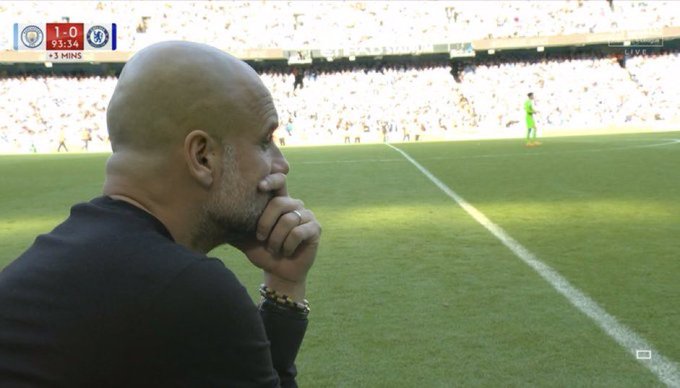 Manchester City manager Pep Guardiola watches from the sideline during a match, with the scoreboard showing Chelsea 1-0 at 93:34 minutes plus 3 added.