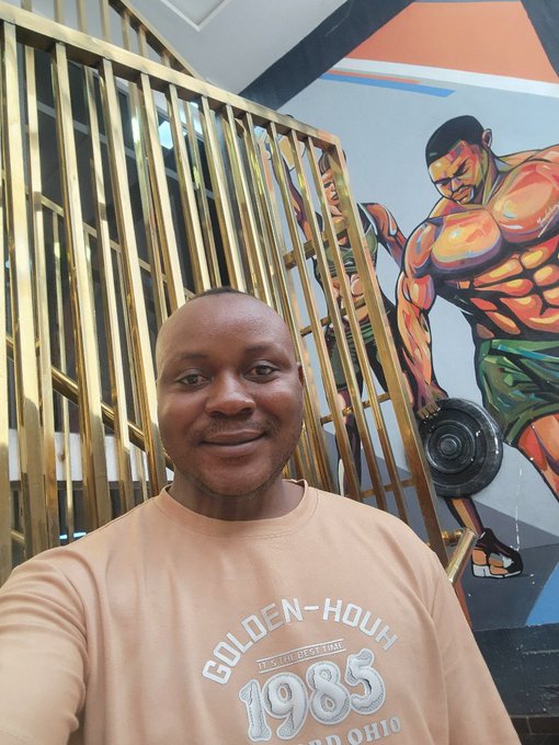 A man wearing a tan "GOLDEN-HOUR 1985" sweatshirt takes a selfie in front of a colorful gym-themed mural featuring bodybuilders and gold-colored bars.